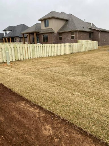 A grassy yard with a wooden fence surrounding a house against a cloudy sky.